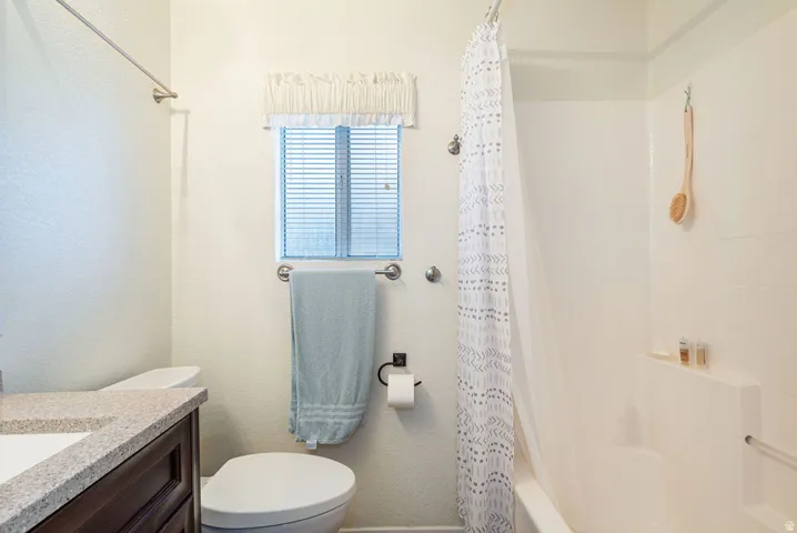 Bathroom featuring a window with blinds, a toilet, a shower-over-tub combination with a built-in shelf, and a vanity with a countertop