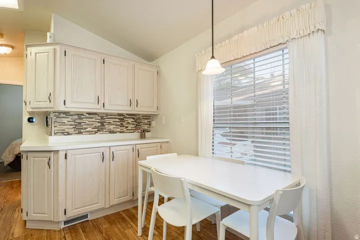 Wood-finish flooring, white cabinetry with dark hardware, patterned tile backsplash, window with blinds and valance, and a pendant light fixture