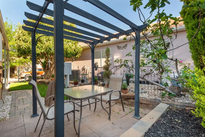 Paved patio area featuring a dark-toned pergola, stone paver flooring, raised garden beds, and mature plantings