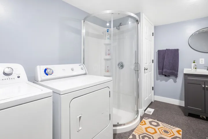 Dedicated laundry room featuring side-by-side washer and dryer appliances, a curved glass shower enclosure with chrome fixtures, dark wood-finish vanity with integrated sink, and warm-toned wood-finish flooring throughout