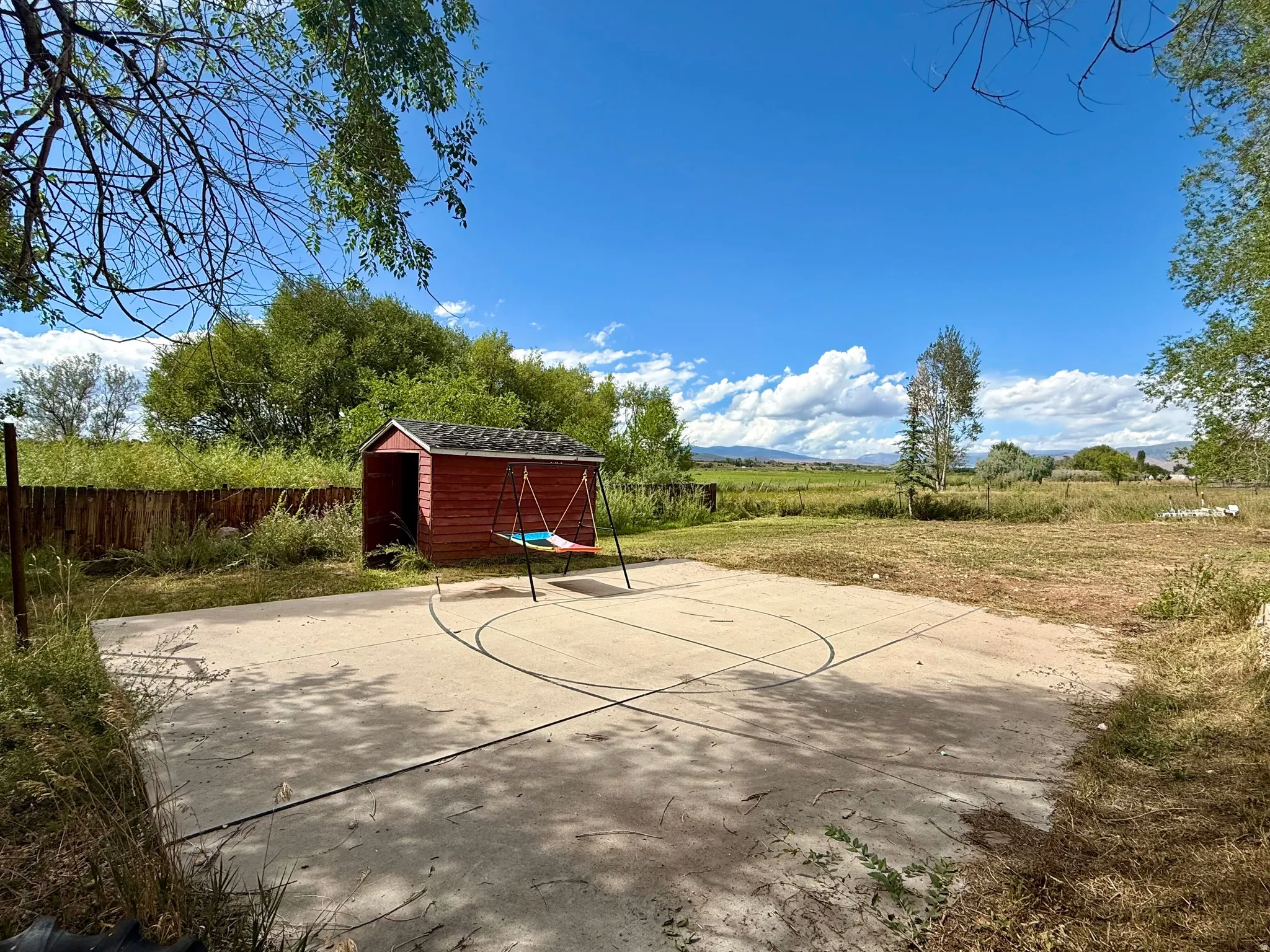 Concrete sport court with painted lines, red wood storage shed, wood fencing, and mature trees