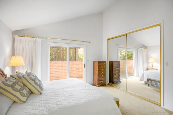 Bright primary bedroom featuring large glass sliding doors with integrated blinds, mirrored closet doors with brass framing, wood-finish flooring, and recessed ceiling lighting