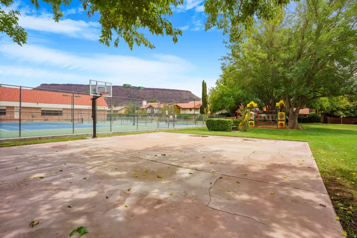 Outdoor basketball court with a chain-link fence, tennis courts, and a playground structure on a grassy area