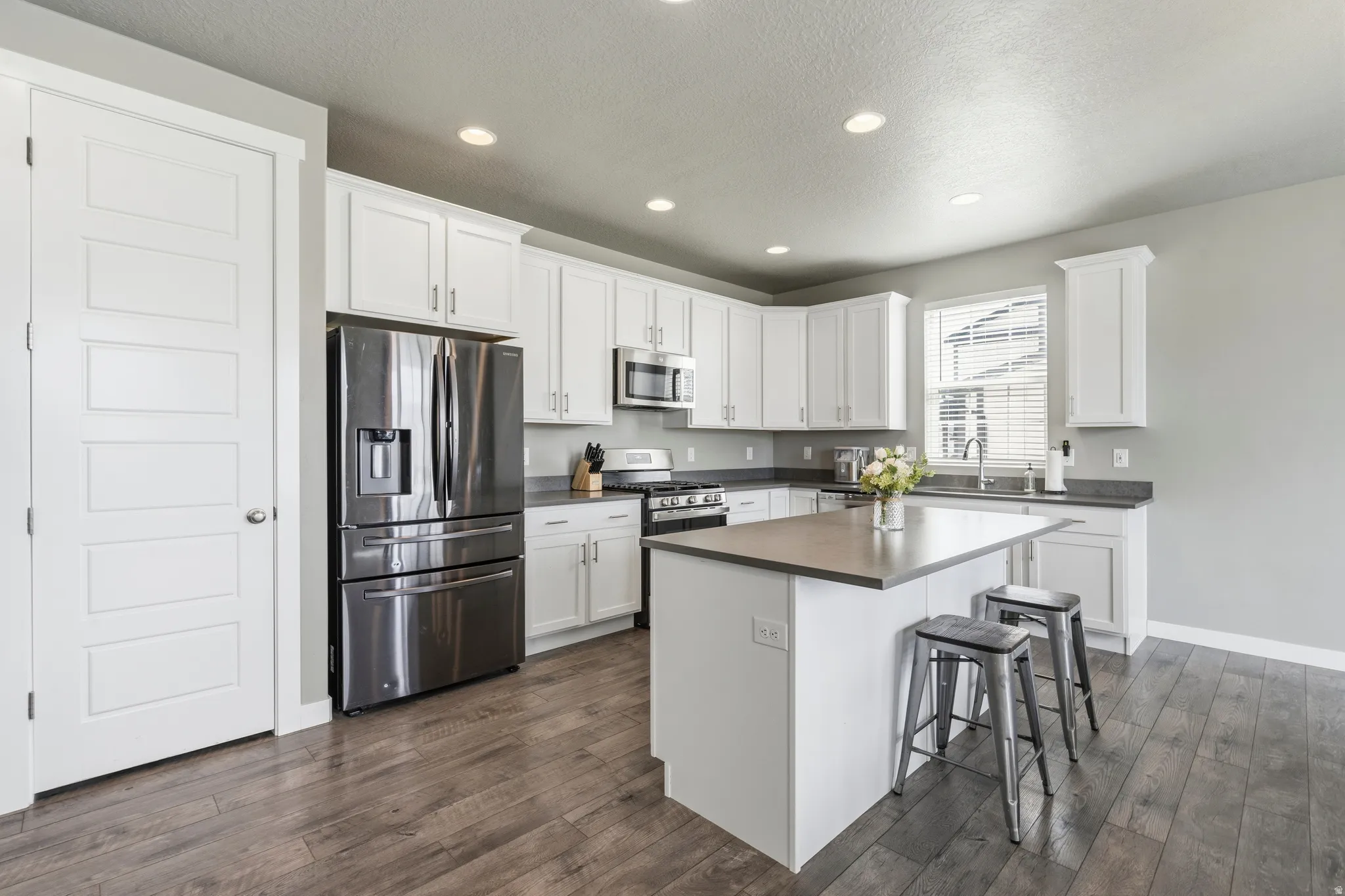 White shaker cabinetry, stainless steel appliances, central island with seating, wood-finish flooring, and recessed lighting
