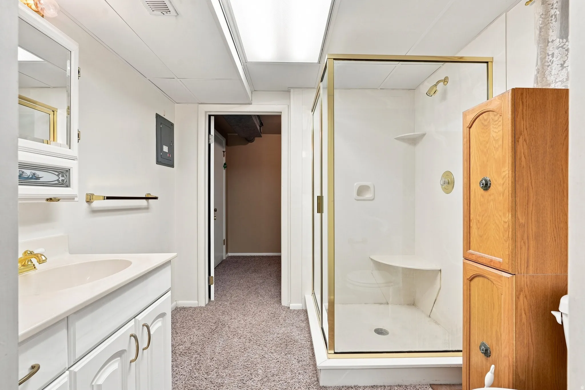 Bathroom featuring a white vanity with integrated sink, gold-tone fixtures, a glass-enclosed shower with gold-tone frame, and a wood-finish storage cabinet