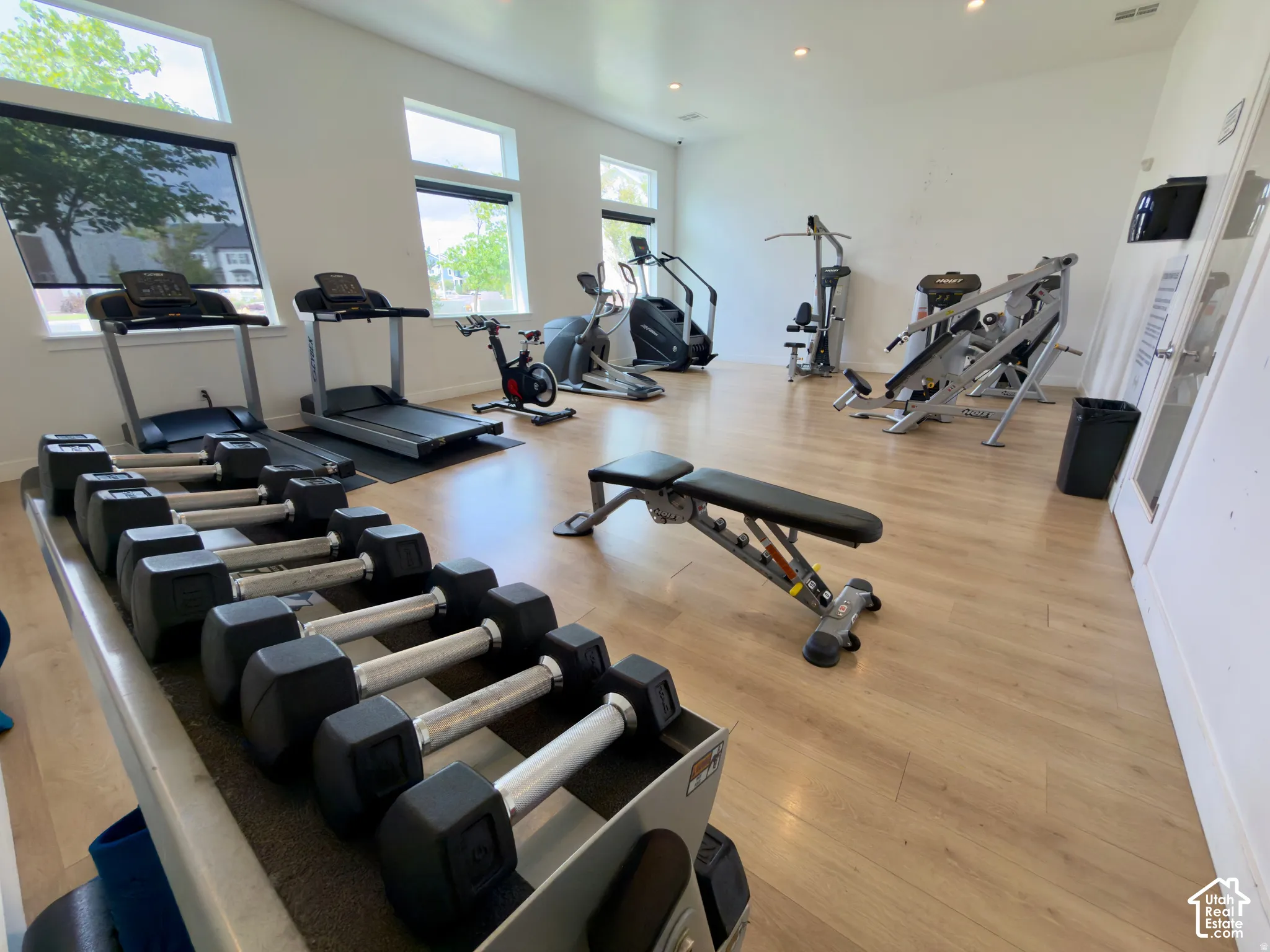 Dedicated fitness area featuring light wood-finish flooring, multiple windows with roller shades, and recessed lighting