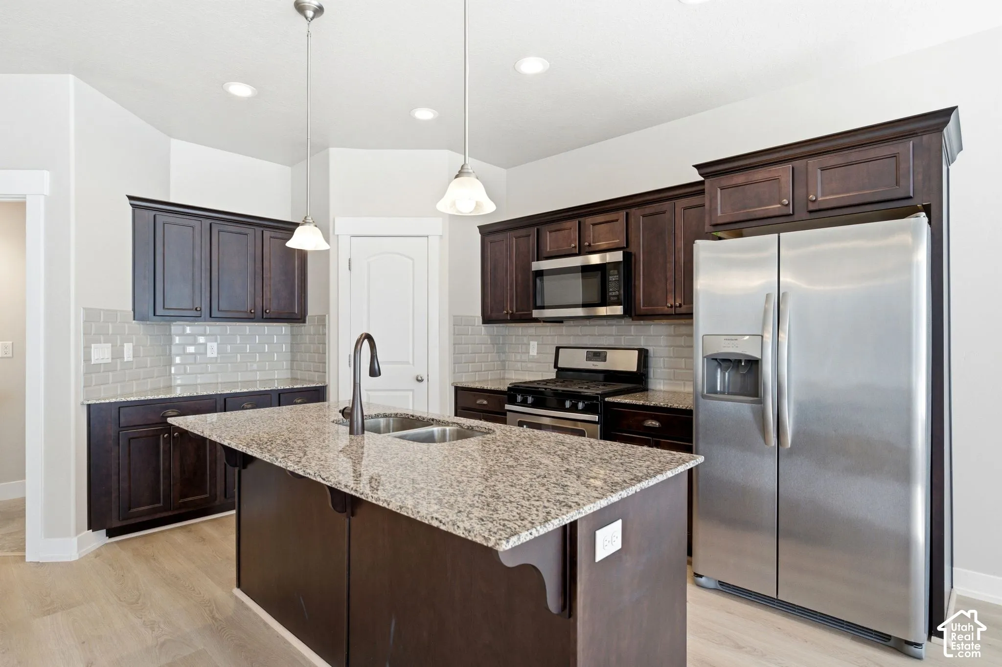 Kitchen featuring a central island with a dual-basin sink, dark wood-finish cabinetry, light-toned countertops, and a subway tile backsplash