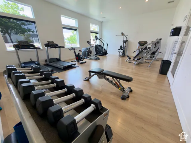 Dedicated fitness area featuring light wood-finish flooring, multiple windows with roller shades, and recessed lighting