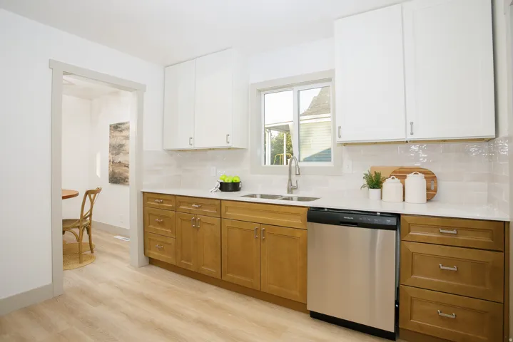 Two-tone cabinetry with white upper cabinets and wood-finish lower cabinets, stainless steel dishwasher, undermount sink with a gooseneck faucet, light-toned backsplash, and white countertops