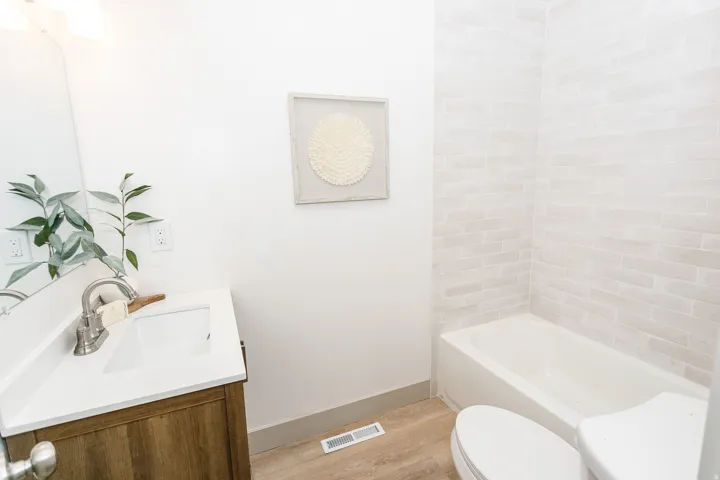 Bathroom featuring a built-in tub with light-toned tiled surround, wood-finish flooring, a vanity with a white countertop, and a wall-mounted light fixture