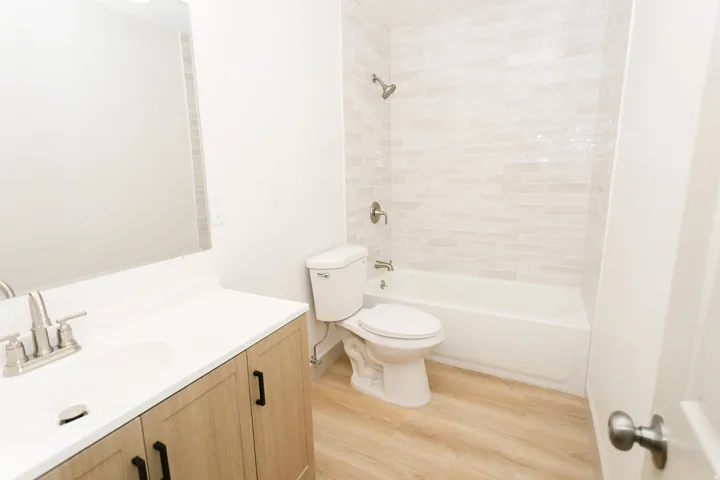 Vanity with a white countertop and wood-finish cabinetry, brushed nickel faucet, large mirror, white bathtub, and light-toned tile surround