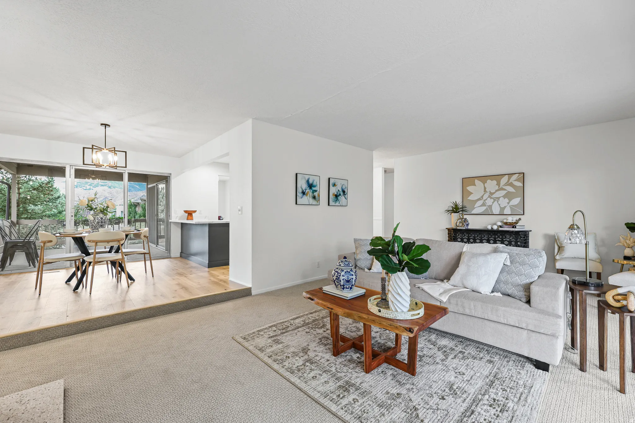 Carpeted living area with abundant natural light, wood-finish flooring in the dining space, a modern dining chandelier, and a built-in kitchen peninsula with a light-toned countertop