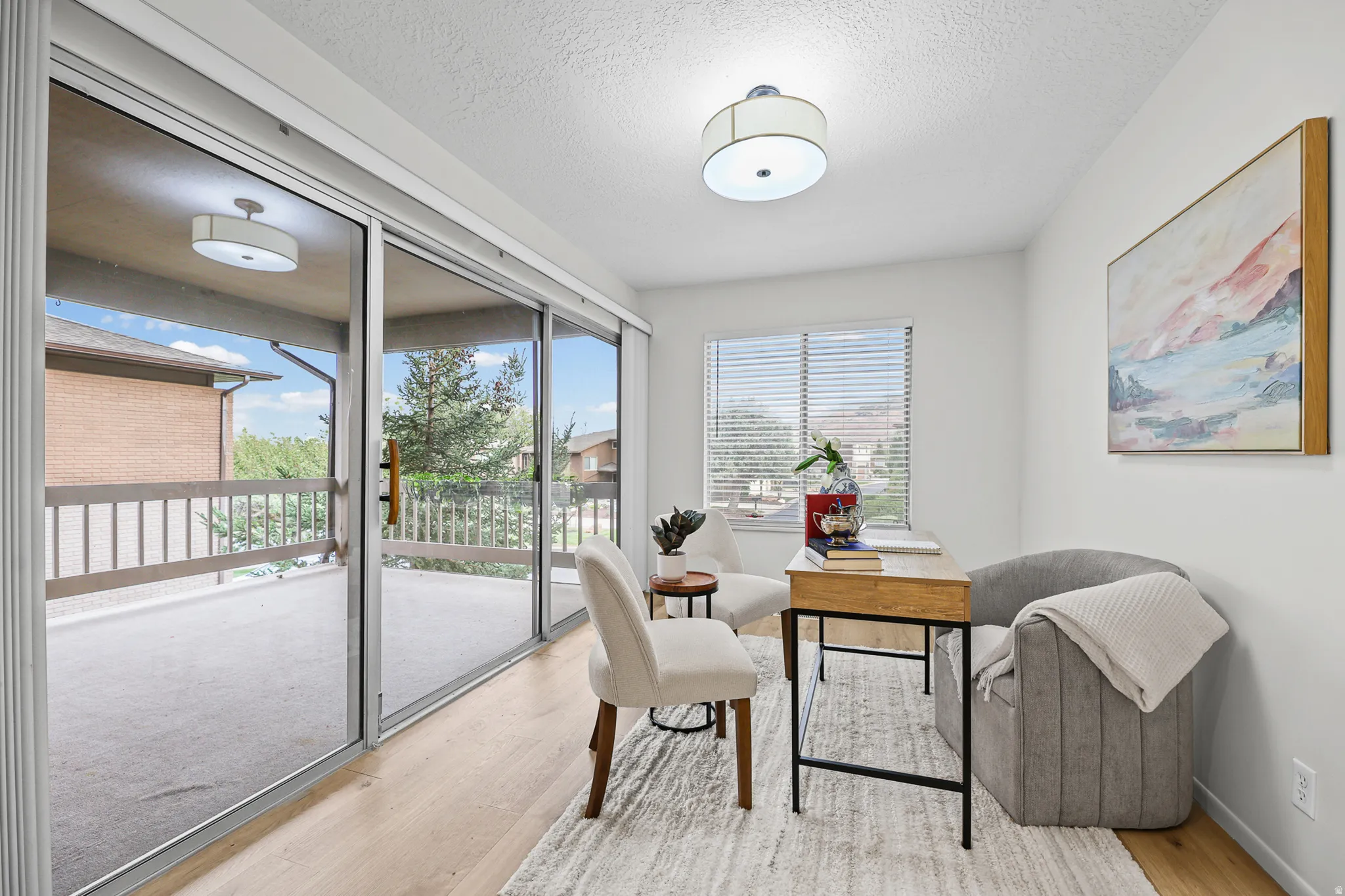 Bright corner room with sliding glass doors opening to a spacious balcony, wood-finish flooring, and abundant natural light from multiple windows