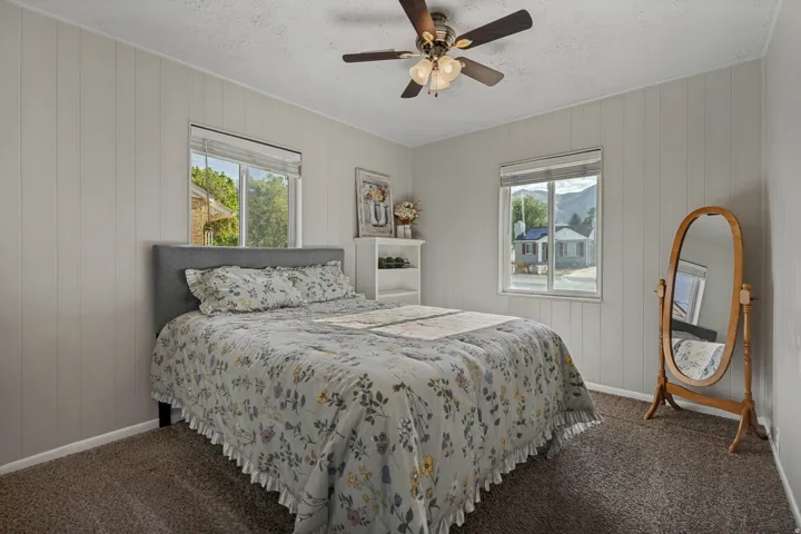 Textured wall panels, ceiling fan with integrated lighting, two windows with blinds, and neutral carpeting