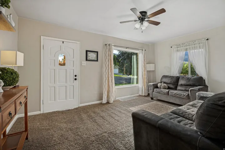 Carpeted living area featuring a multi-pane front entry door, a large picture window, and a ceiling fan with light fixture
