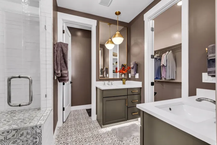 Dual vanity sinks with white countertops, decorative floor tiling, walk-in shower with bench, and integrated closet storage
