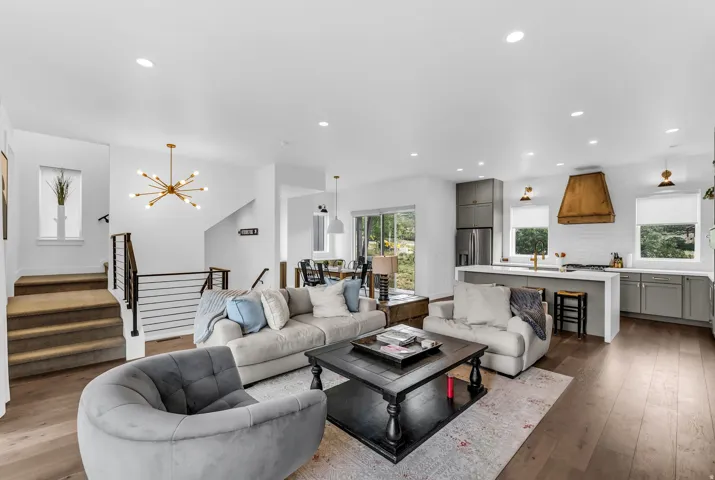 Spacious living area featuring wood-finish flooring, a modern kitchen with a large island and wood-paneled range hood, recessed lighting, and a contemporary chandelier