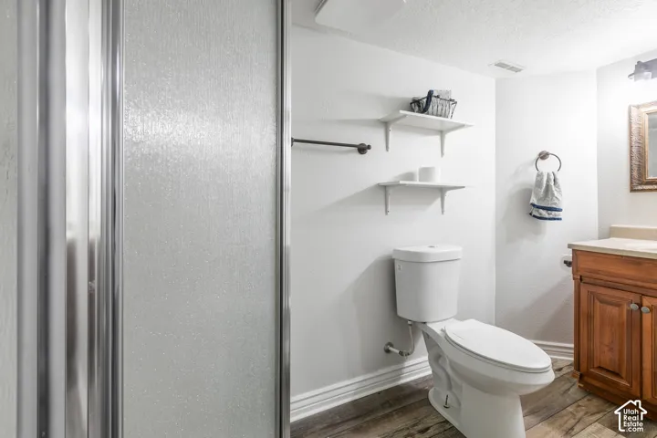 Frosted glass shower enclosure, wood-finish flooring, white walls, wood vanity with a light-toned countertop, and wall-mounted shelving