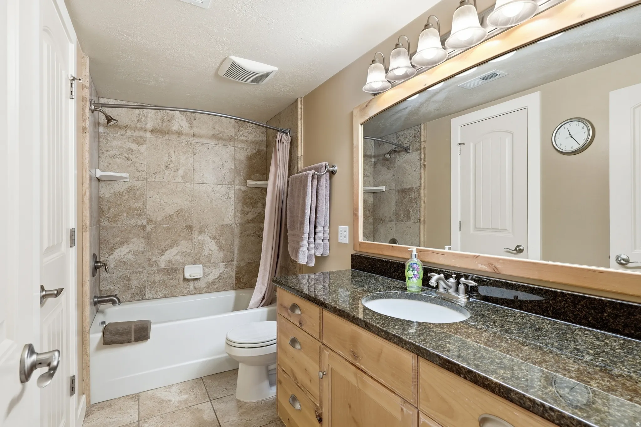 Vanity with integrated sink and dark speckled countertop, light wood-finish cabinetry, frameless mirror with wood trim, and four-light vanity fixture