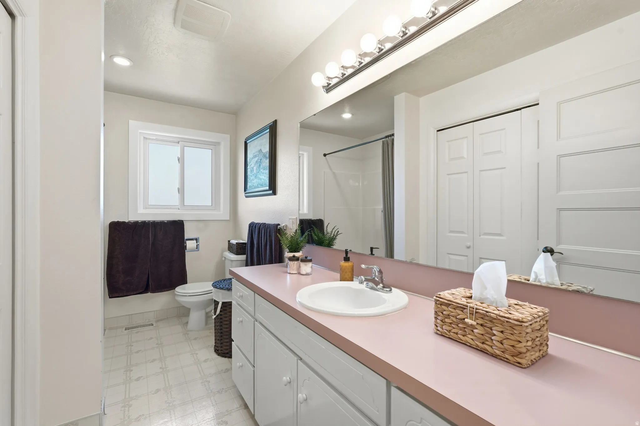 Bathroom featuring a single vanity with an integrated sink, wall-mounted mirror, overhead vanity lighting, and a sliding window