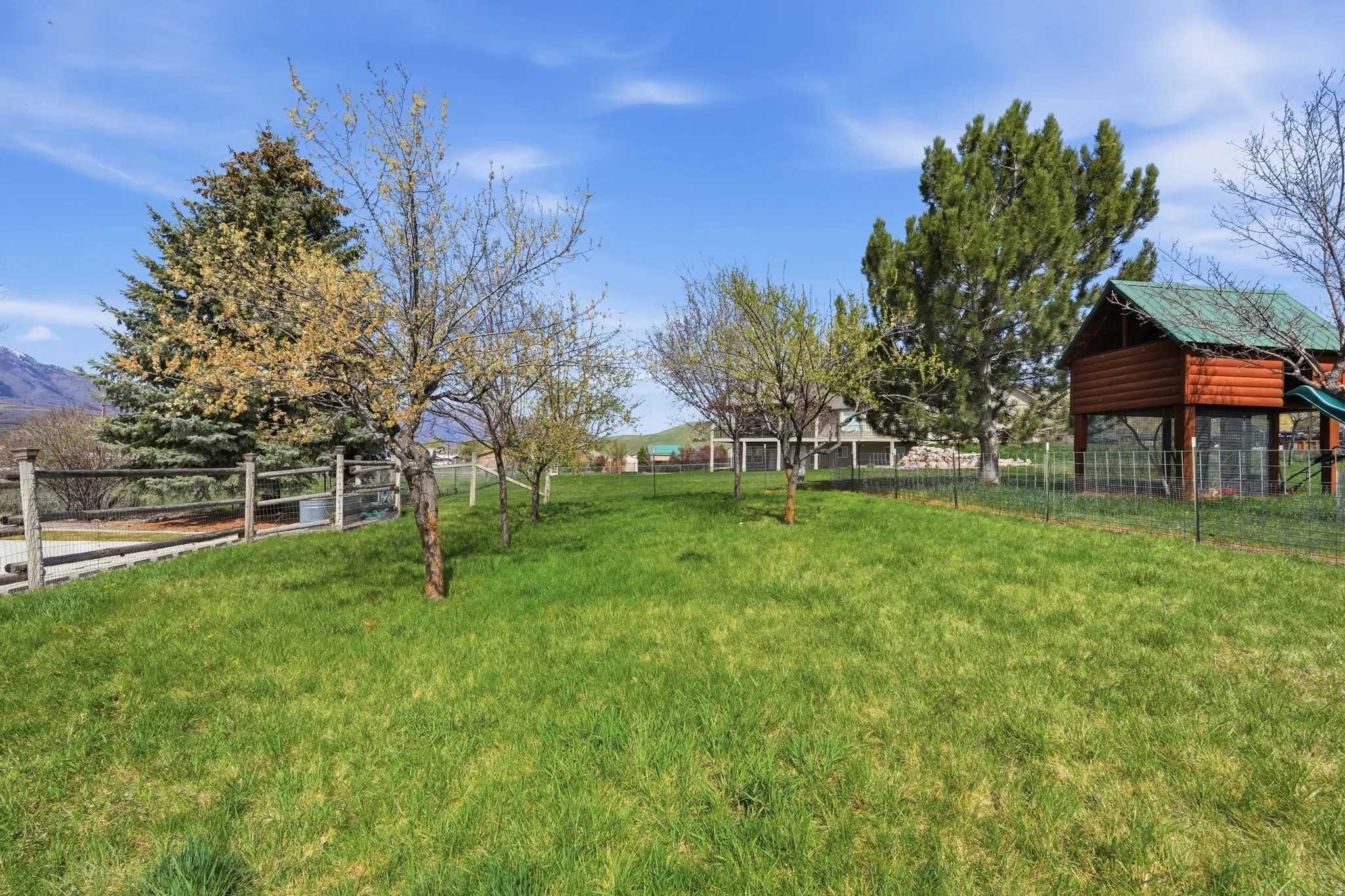 Expansive grassy yard with mature trees, a rustic split-rail fence, and a log-style outbuilding with a green roof