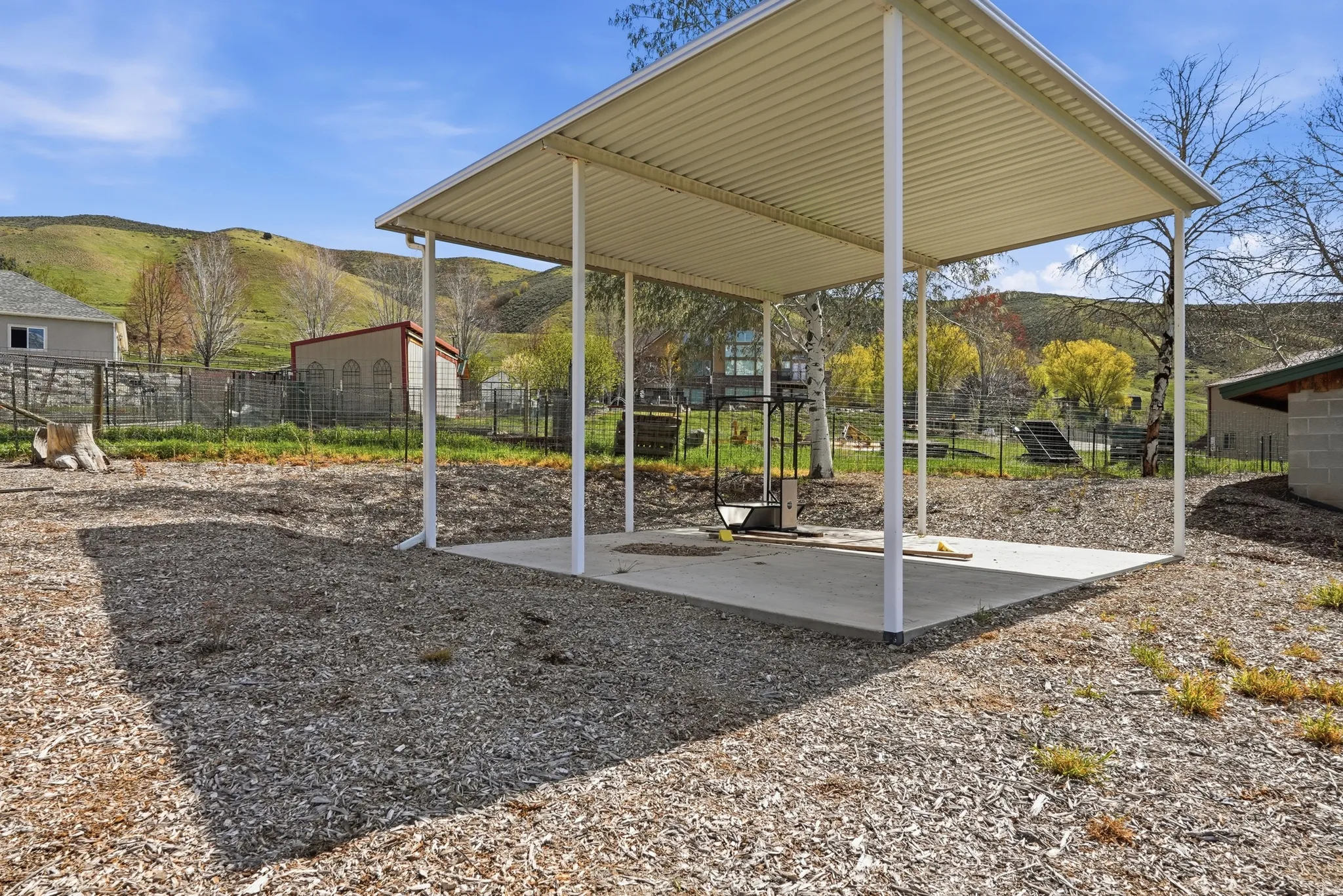 Spacious covered patio with white metal support columns and gable roof, situated on gravel ground with mature trees and rolling hillside backdrop