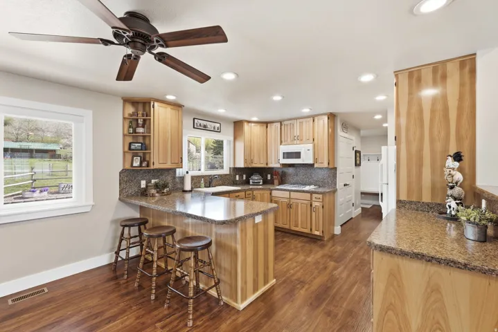 Wood-finish flooring, recessed lighting, and a ceiling fan illuminate this kitchen featuring light wood cabinetry, stone countertops, and a built-in breakfast bar