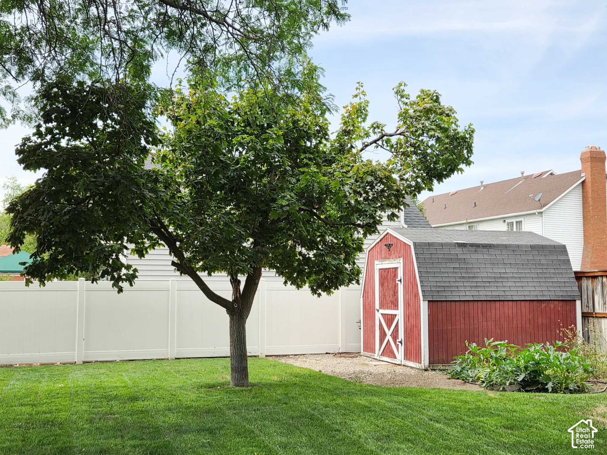 Expansive backyard featuring a manicured lawn, established tree, white vinyl fencing, and a red storage shed with a gambrel roof