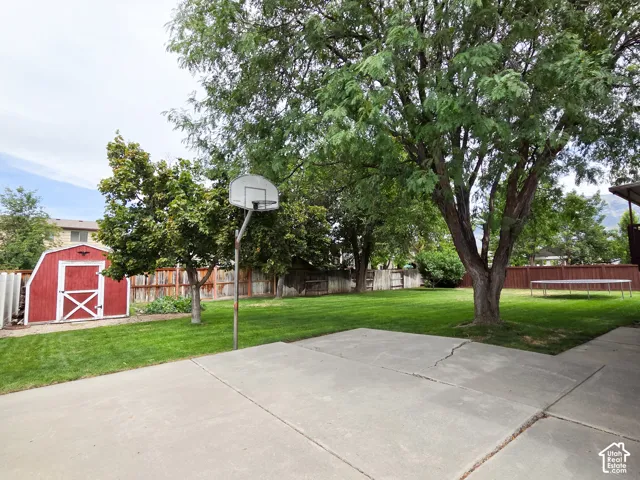 Expansive backyard featuring a red storage shed, mature trees, a concrete patio, and a basketball hoop