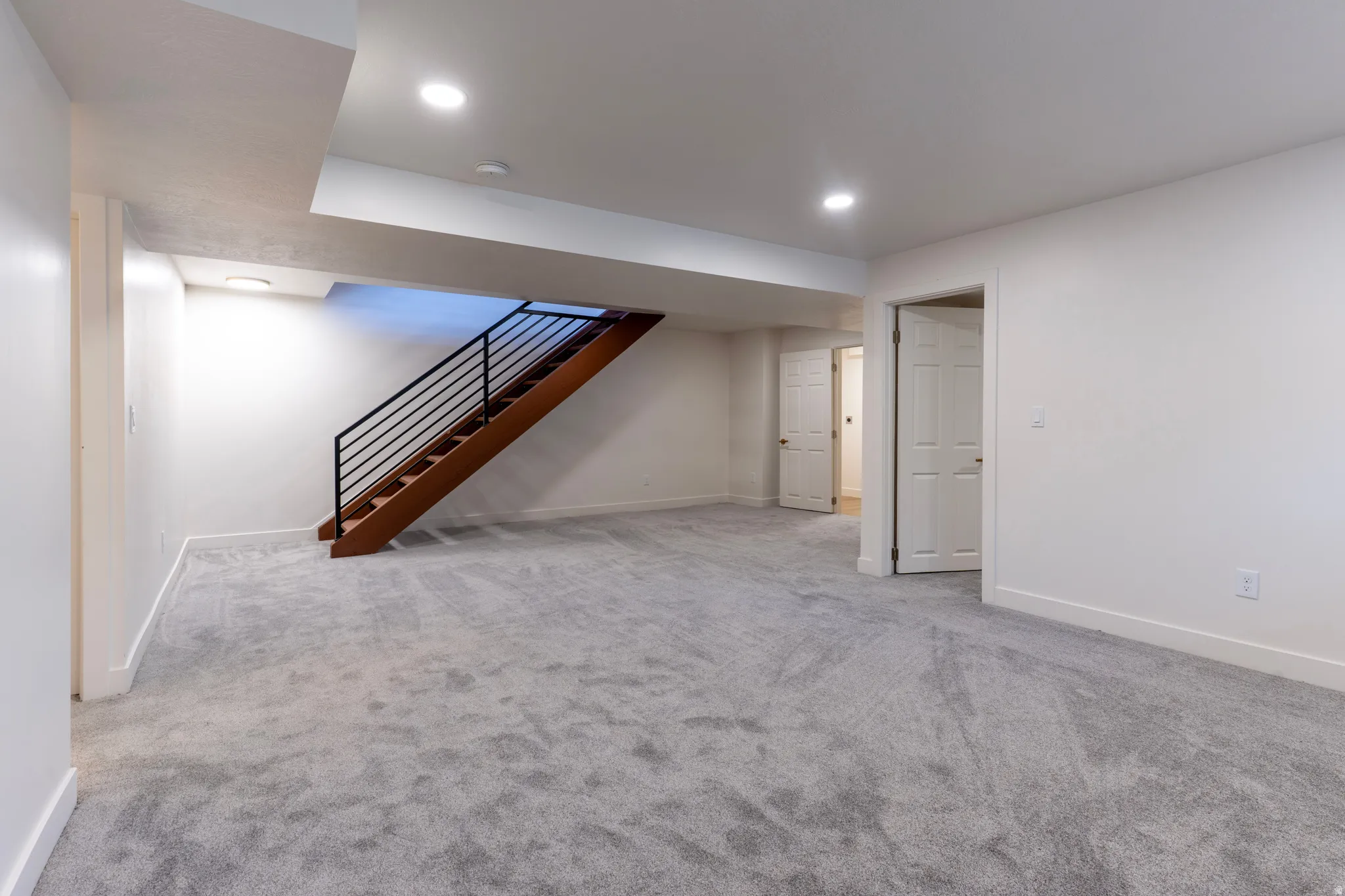 Spacious room with light gray carpeting, recessed ceiling lighting, and a wood staircase with black metal railing