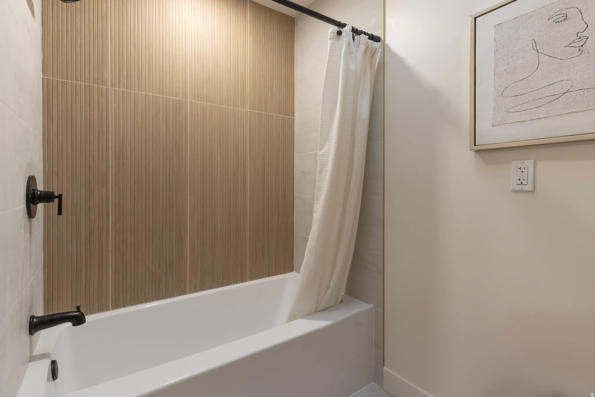 Bathtub with matte black fixtures, textured beige wall tile, and light-toned wall tile