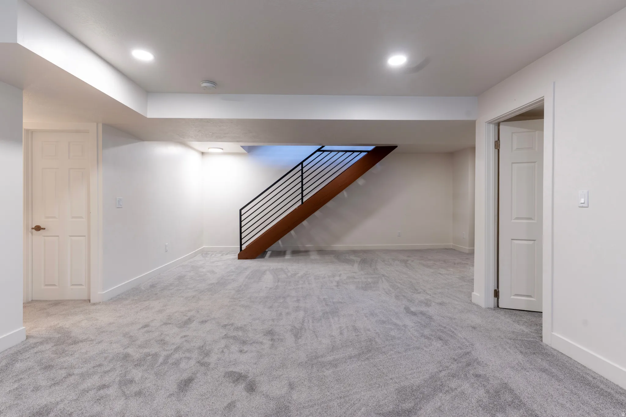 Spacious room with plush gray carpeting, recessed ceiling lighting, and a modern staircase featuring wood-finish treads and black metal railings