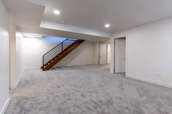 Spacious room with light gray carpeting, recessed ceiling lighting, and a wood staircase with black metal railing