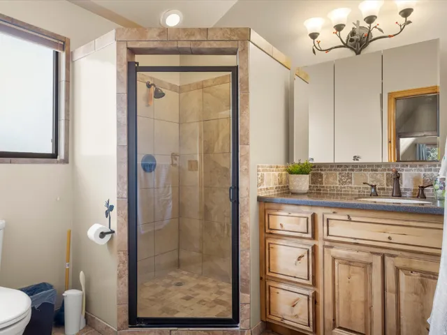 Walk-in shower with tiled surround and glass enclosure, wood-finish vanity with countertop, decorative tile backsplash, and wall-mounted vanity light