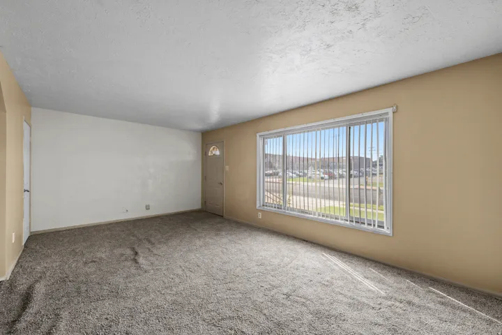 Carpeted flooring, neutral wall tones, textured ceiling, and a large window with vertical blinds