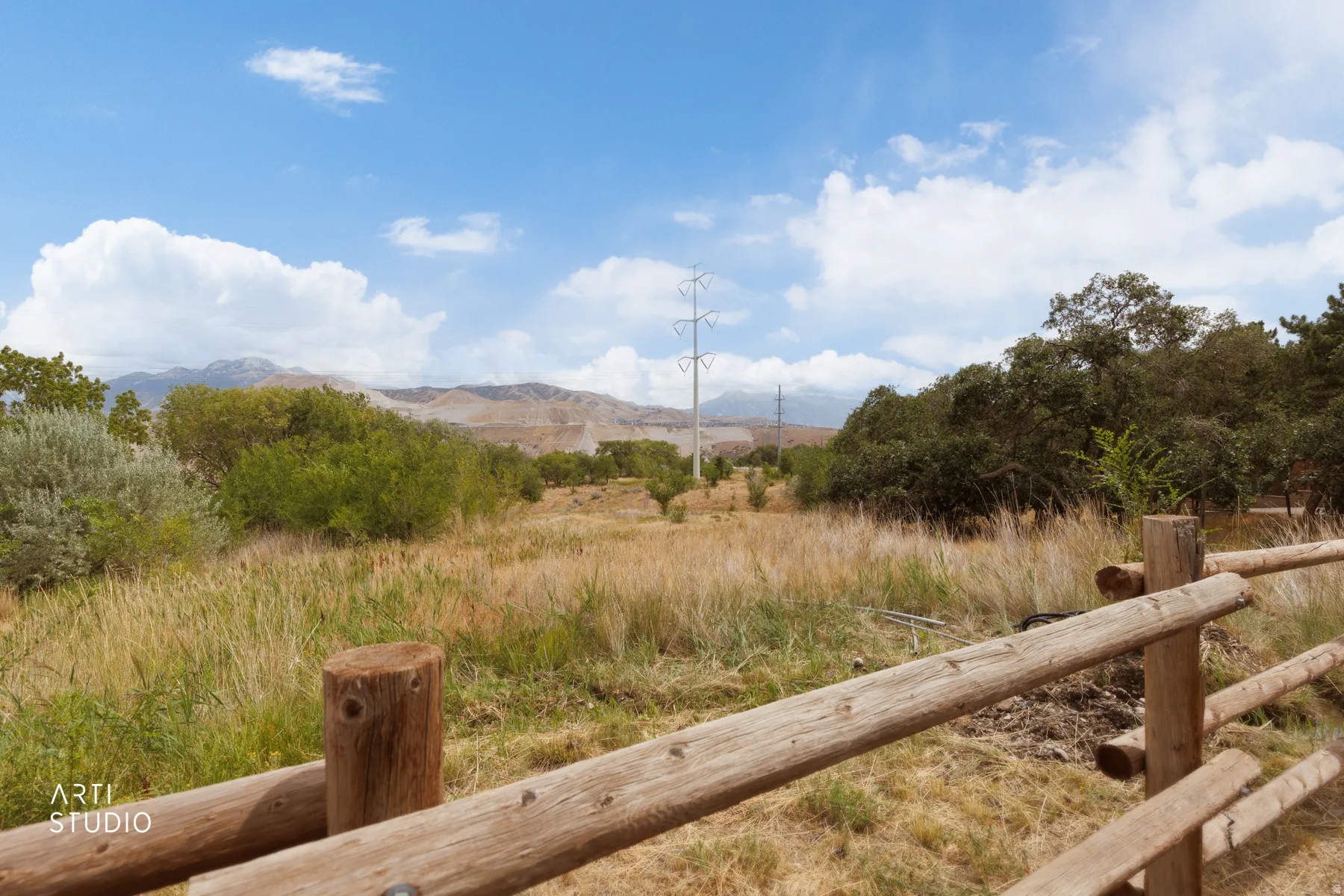 Wooden post-and-rail fencing, expansive natural landscape, distant mountains, mature trees, and clear blue sky with clouds