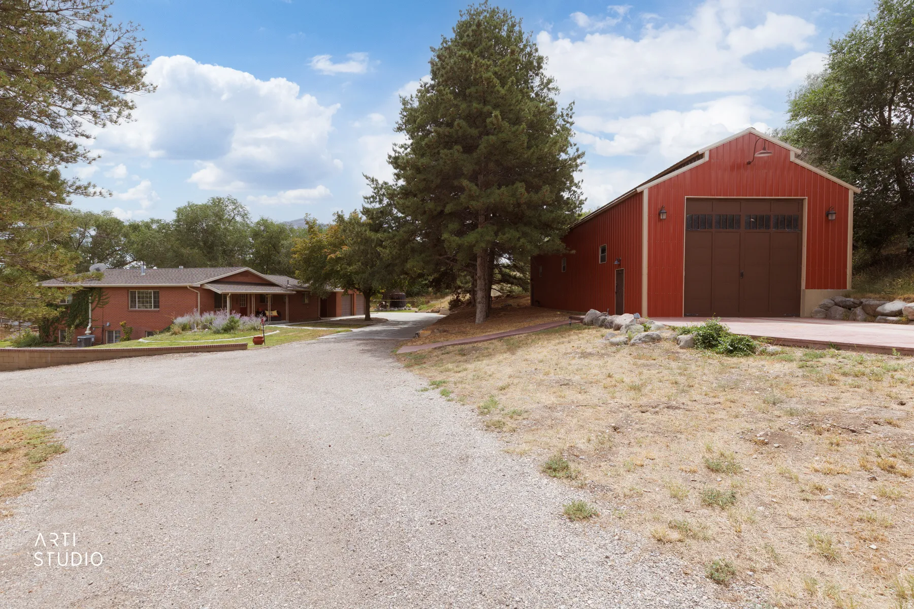 Gravel driveway leading to a red barn-style auxiliary structure with a large garage door, and a brick residence with a covered front porch and multi-pane windows