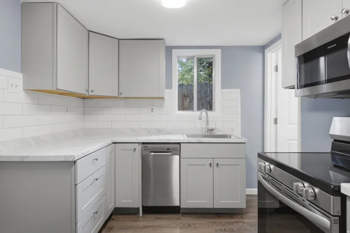 L-shaped kitchen with light gray cabinetry, white subway tile backsplash, stainless steel appliances, and wood-finish flooring