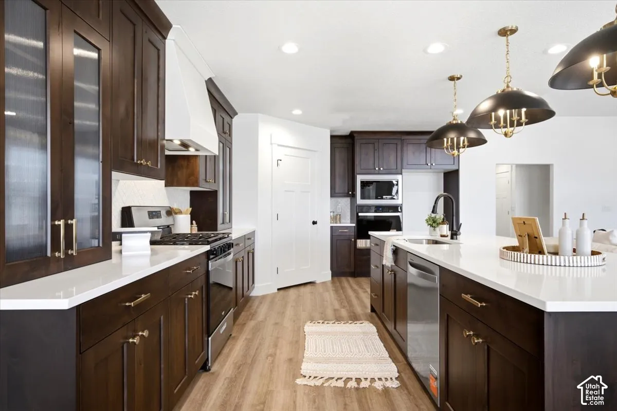 Kitchen featuring dark wood cabinetry, white countertops, stainless steel appliances, a center island with sink, and recessed and pendant lighting