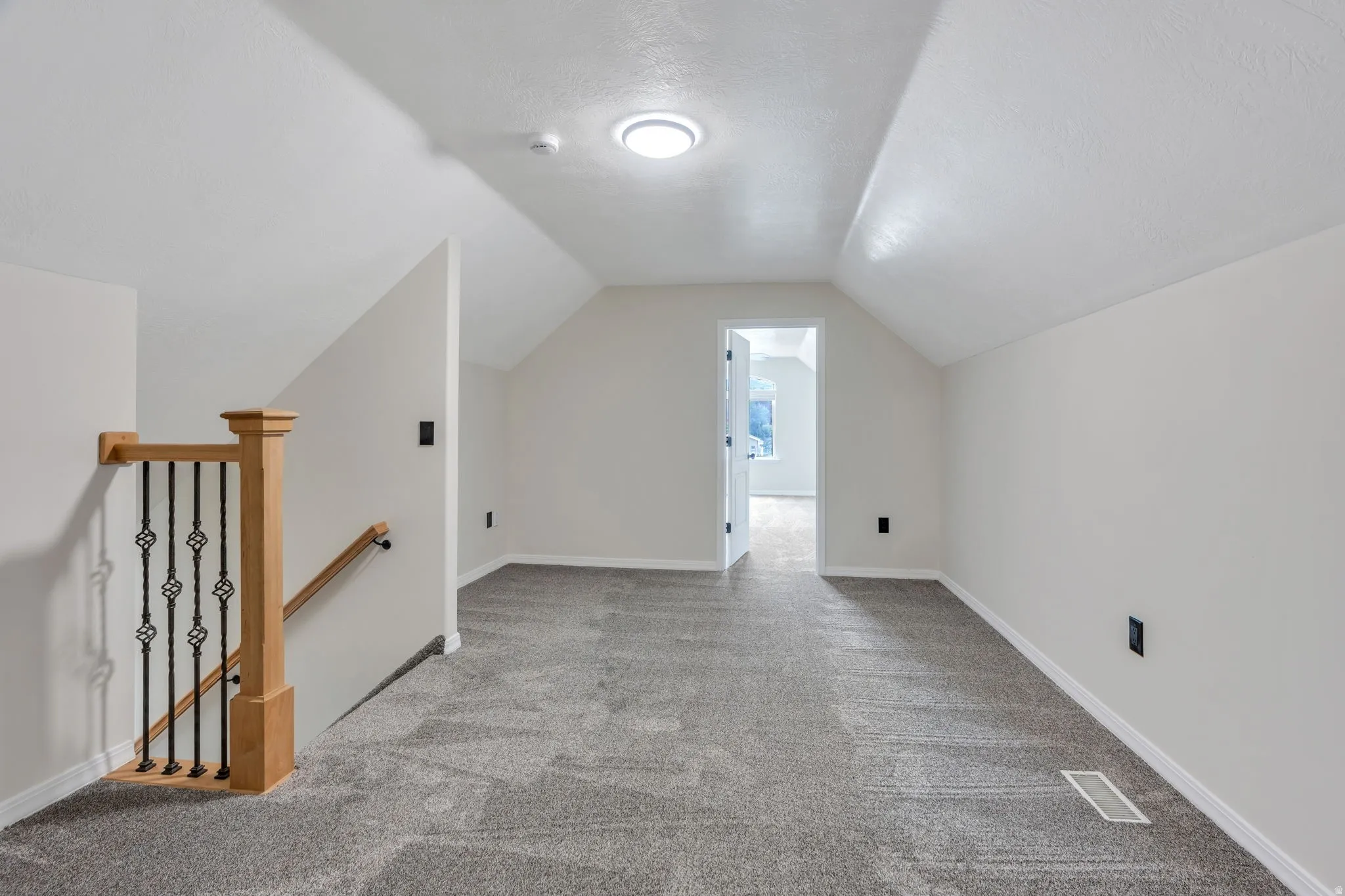Attic space with sloped ceilings, gray carpeting, and a wooden handrail with wrought iron balusters