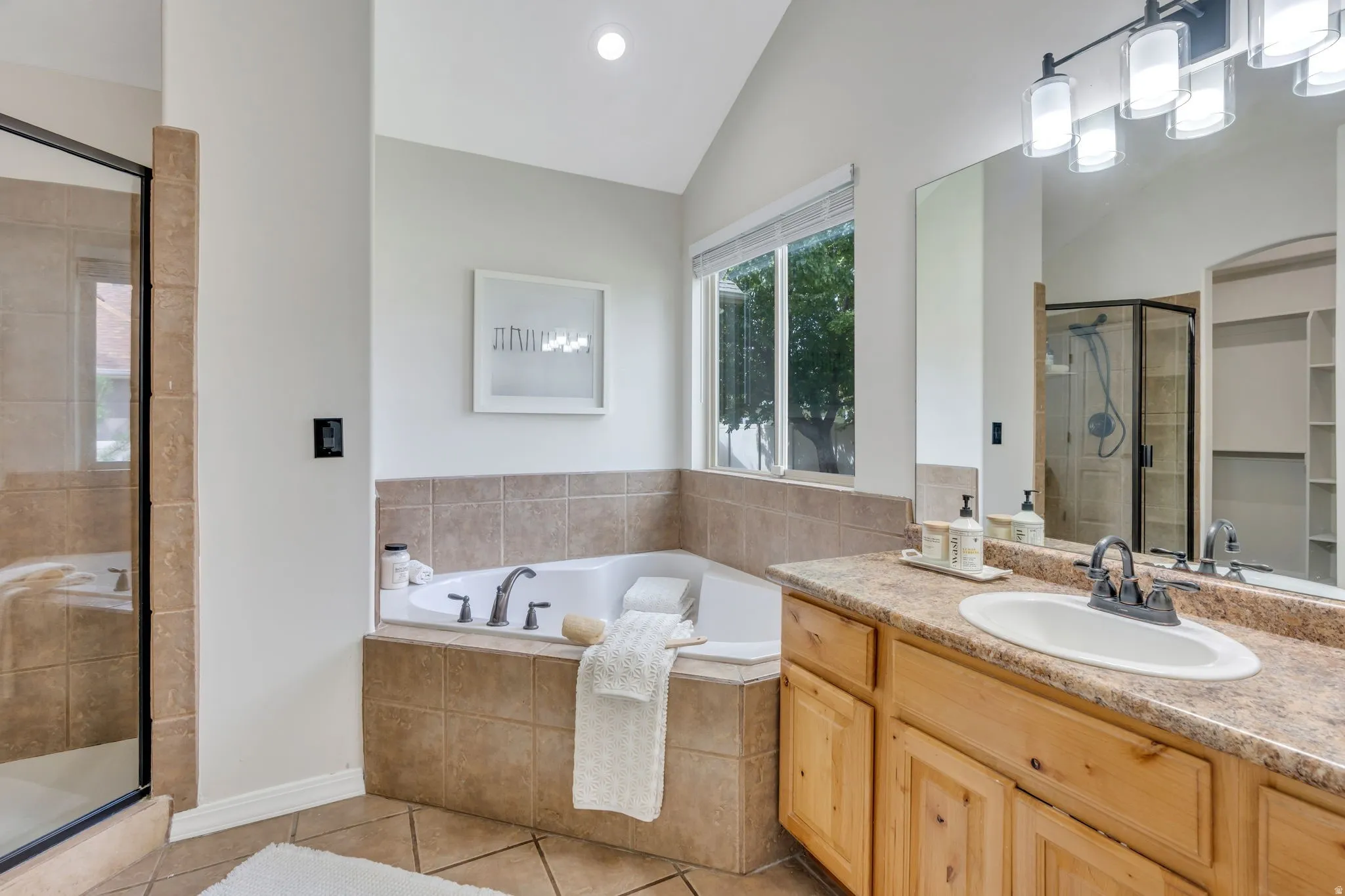 Alcove soaking tub with tiled surround, separate shower with glass enclosure, single vanity with light wood cabinetry and stone-finish countertop, and recessed shelving