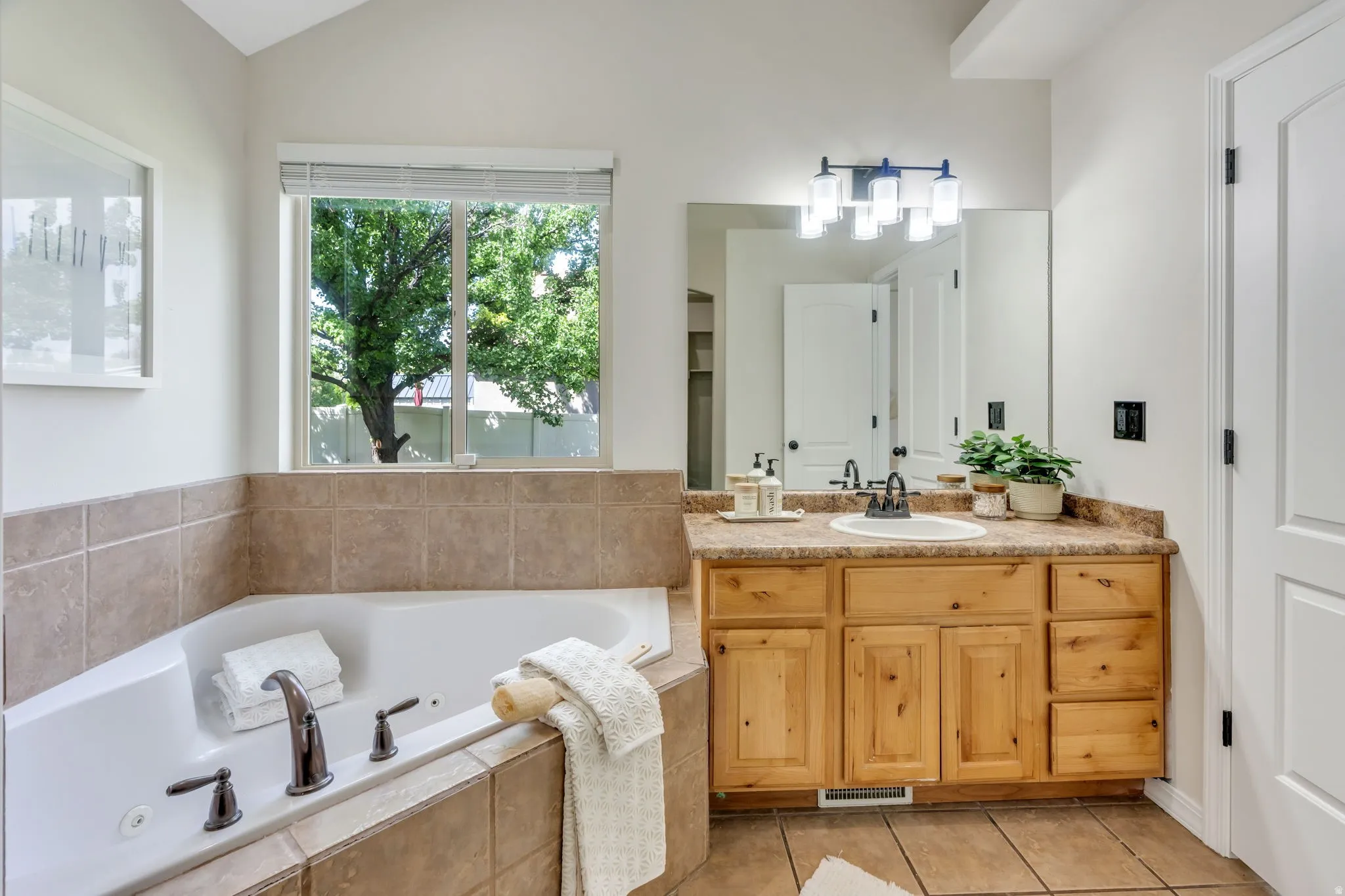 Soaking tub with bronze-finish fixtures, tile surround, wood-finish vanity with an integrated sink, and a wall-mounted light fixture