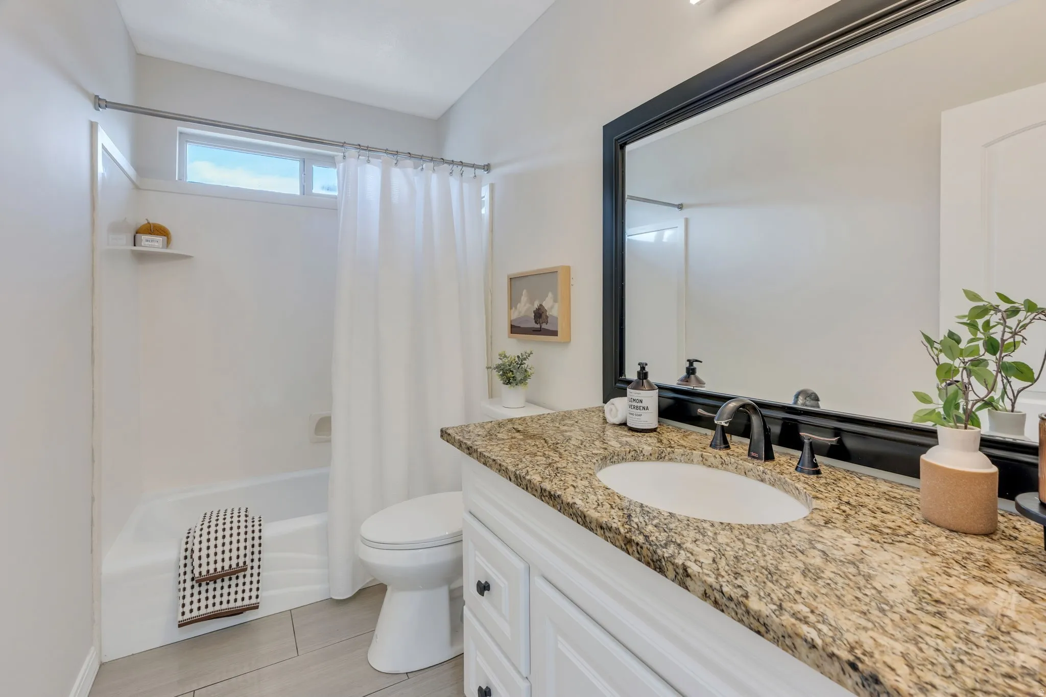 Granite-style countertop with integrated sink, black framed mirror, white vanity cabinetry with black hardware, black-finish faucet, and rectangular floor tiles