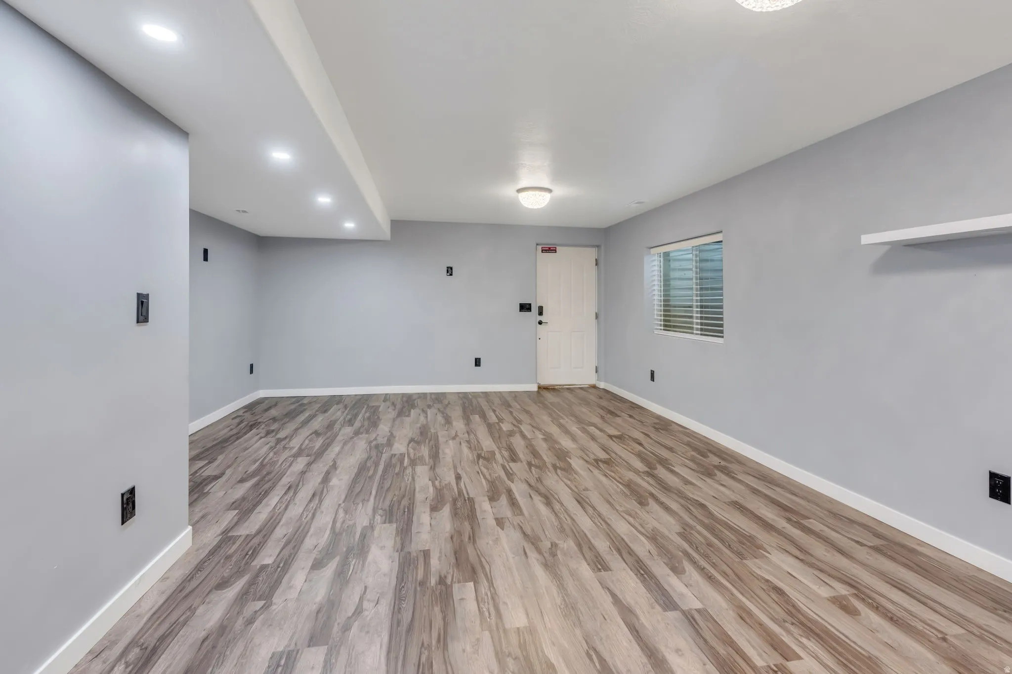 Recessed ceiling lighting, wood-finish flooring, light gray wall paint, a white door, and a horizontal window blind