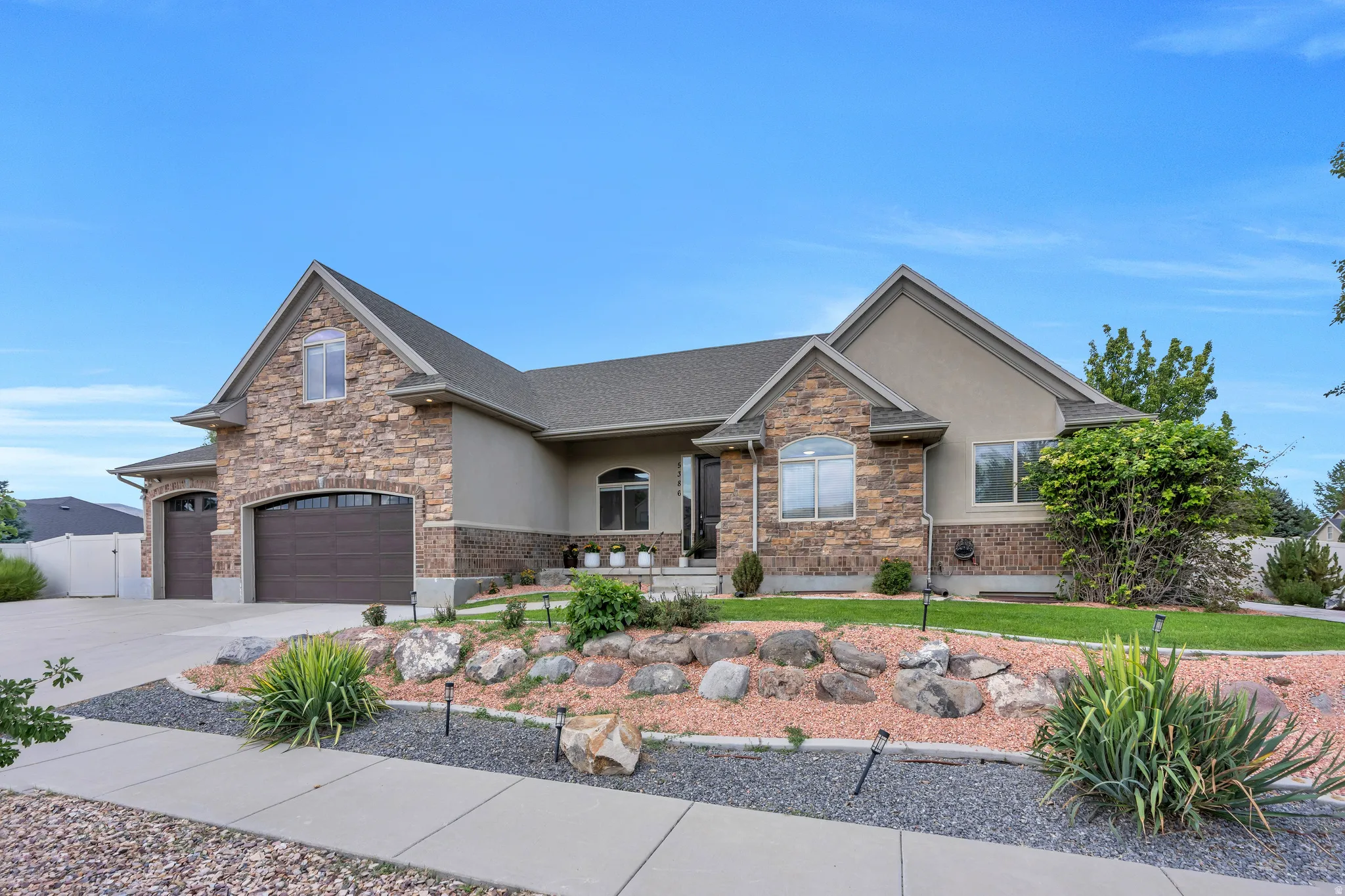 Stone and stucco exterior, arched garage doors, established landscaping with rock features, concrete driveway, and a paved walkway