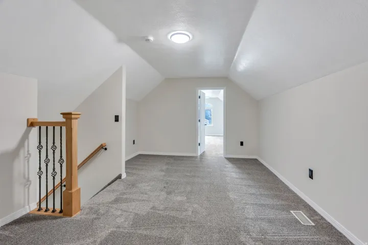 Attic space with sloped ceilings, gray carpeting, and a wooden handrail with wrought iron balusters