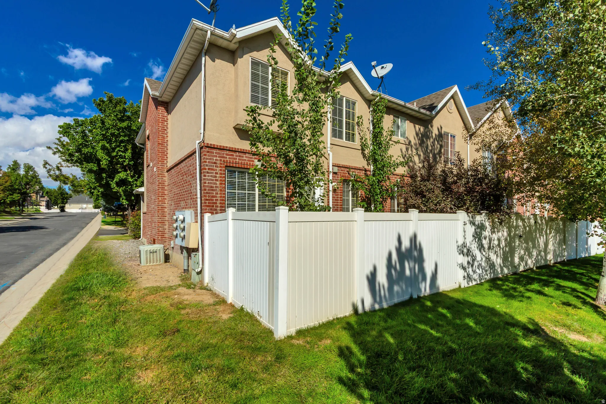 Townhome exterior featuring tan stucco, brick accents, multiple windows, a white privacy fence, and a grassy lawn