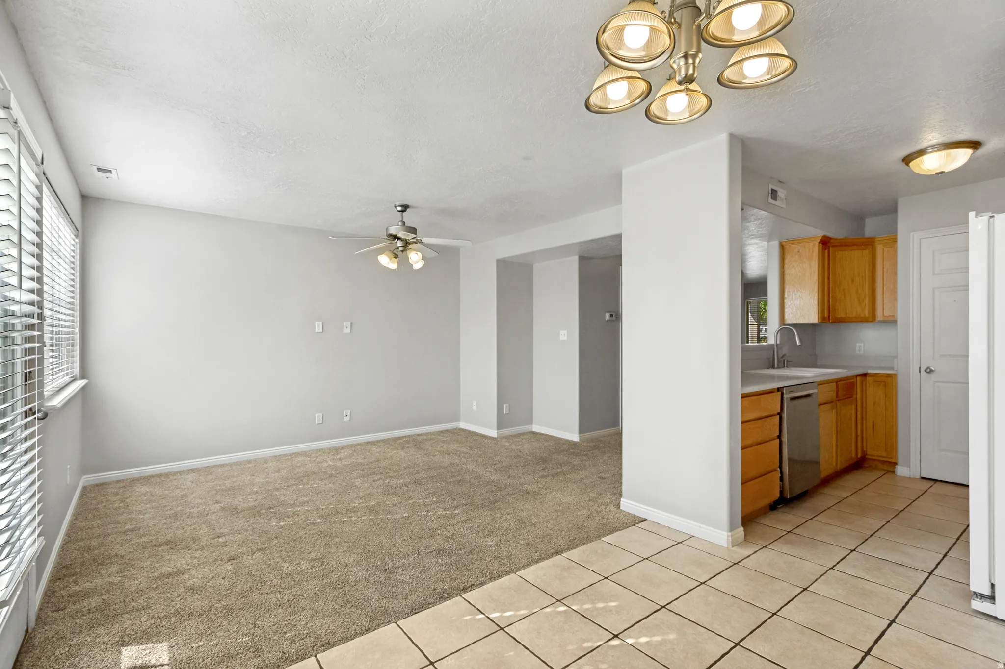 Carpeted living area with window blinds and a ceiling fan, opening to a kitchen with wood cabinetry, tile flooring, and stainless steel appliances