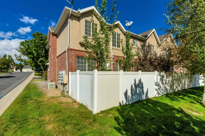 Townhome exterior featuring tan stucco, brick accents, multiple windows, a white privacy fence, and a grassy lawn