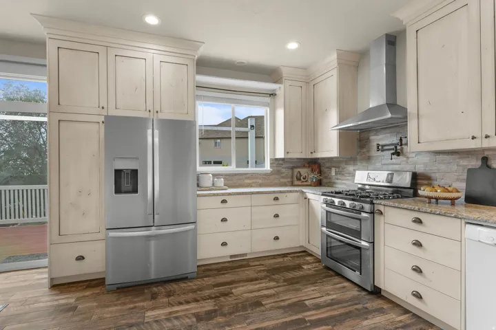 Kitchen featuring wood-finish flooring, light-toned cabinetry, stainless steel appliances, stone backsplash, and granite countertops